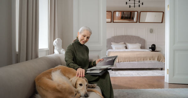 Senior woman reading on a sofa while petting her greyhound dog in a cozy living room.