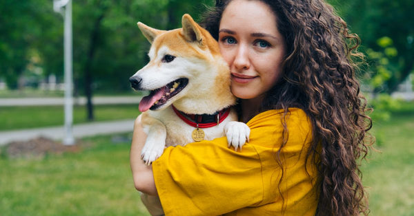 A young woman lovingly holds her Shiba Inu dog in a sunlit park setting.