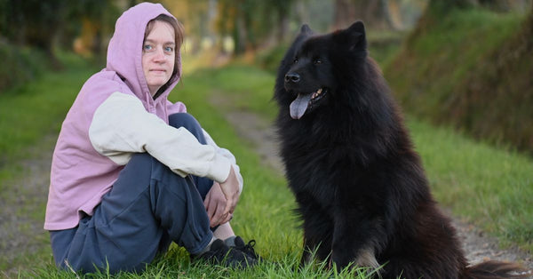 A young woman in a hoodie sitting with a large black dog on a grassy path.