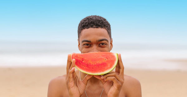 A young man holds a watermelon slice at the beach, capturing a fun summer moment.